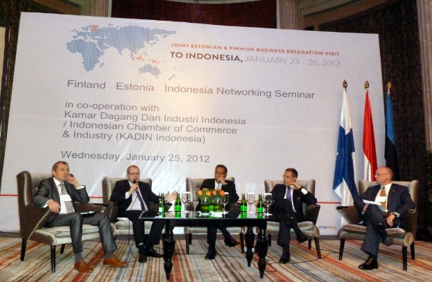 Eine Konferenzbühne mit Menschen, Stühlen, einem Mikrofon, einem Tisch, Flaschen, Blumen, Gläsern und anderen Gegenständen, mit Flaggen und einem Banner in der Mitte und einem Gebäude mit Vorhängen im Hintergrund.