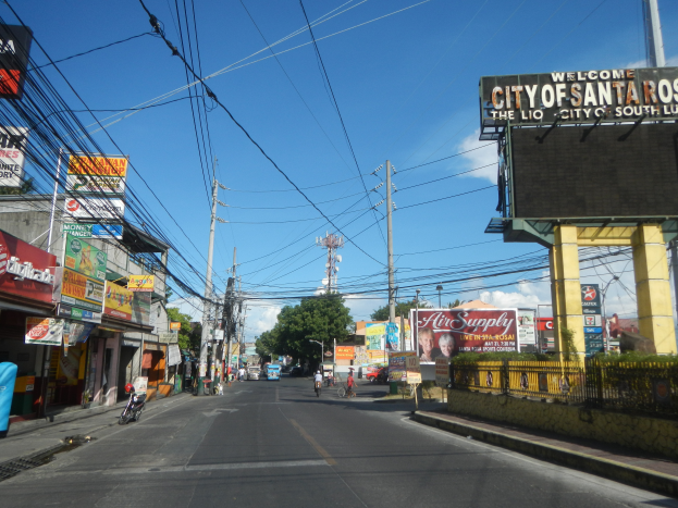 Eine belebte Stadtstraße mit Fahrzeugen, Radfahrern, Gebäuden und einem Schild auf der rechten Seite, das "Willkommen in Santa Rosa" lautet, mit einem bewölkten Himmel im Hintergrund.