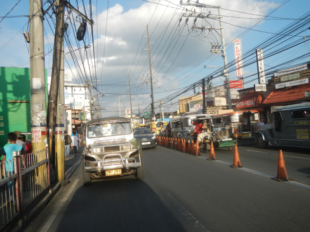 Eine belebte Stra├če mit Fahrzeugen, Absperrgittern, Passanten, Stromleitungen, Geb├Ąuden und einem bew├Âlktem Himmel.