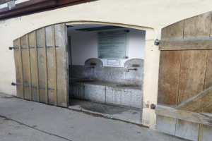 Ein altes Gebäude mit einer hölzernen Tür und einem Schild, das einen Brunnen mit zwei Hähnen und eine Tafel zeigt, die wahrscheinlich ein öffentliches WC anzeigt, mit dem Boden unten.