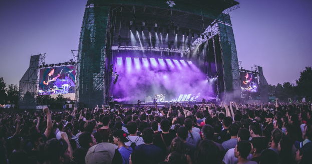 Eine große Menschenmenge auf einem Musikfestival schaut Künstlern auf einer hell erleuchteten Bühne mit Bildschirmen zu, die vor einem Gebäude, Bäumen und einem blauen Himmel aufgebaut ist.