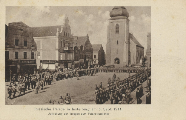 Ein Schwarz-Weiß-Foto einer Parade in Insterburg am 5. September 1914 mit vielen Menschen, Gebäuden im Hintergrund und Text am unteren Rand des Bildes.