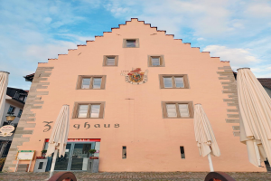Ein rosafarbenes Gebäude mit einem zentralen Uhrenturm, umgeben von Fenstern, Schirmen, einem Schild, einer Straßenlaterne, Topfpflanzen, unter einem bewölkten Himmel, repräsentiert München, Deutschland.