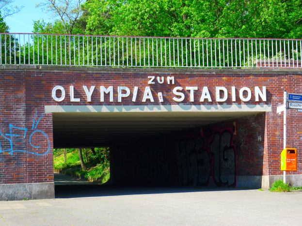 Der Eingang zum Olympiastadion in Berlin, Deutschland, mit einer Brücke mit Text, einem Metallzaun, einem Schild, einer Box, Pflanzen, Gras, einer Baumgruppe und einem bewölkten Himmel.