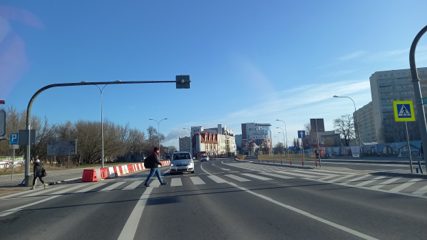 Ein Mann überquert eine Straße an einer Ampel, mit Fahrzeugen, Fußgängern, Verkehrskegeln, Verkehrszeichen, Straßenlaternen, Bäumen, Gebäuden und einem bewölkten Himmel im Hintergrund.