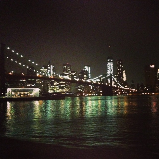 Mehrere Gebäude mit Lichtern, eine Brücke über Wasser und zusätzliche Lichter unter einem Nachthimmel.