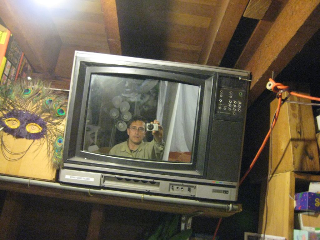 Ein Fernsehbildschirm mit einer Person, die von Pfauenfedern, Büchern und Kartons umgeben ist.