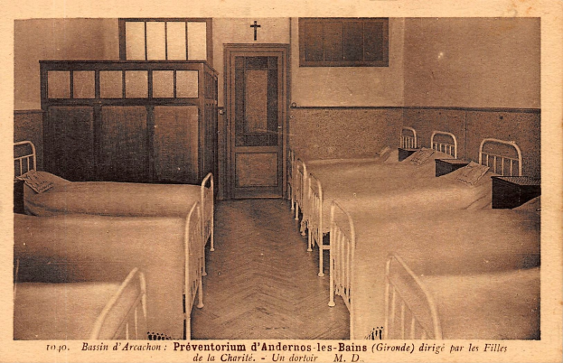 Ein altes Foto eines Krankenhauszimmers mit Betten, Kissen, Tischen, einer Tür, Fenstern und einem Kreuz an der Wand, mit Text unten.