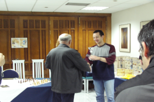 Ein Besprechungsraum mit Menschen, die stehen und sprechen, ein blauer Tisch mit einem Schachbrett, ein Stuhl davor, Schränke im Hintergrund und Fotorahmen an der rechten Wand.