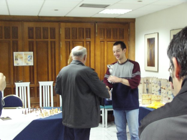 Ein Besprechungsraum mit Menschen, die stehen und sprechen, ein blauer Tisch mit einem Schachbrett, ein Stuhl davor, Schränke im Hintergrund und Fotorahmen an der rechten Wand.