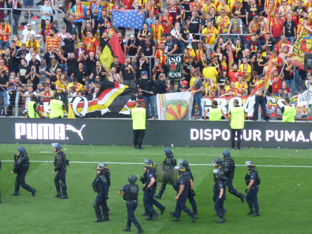 Eine Gruppe von Polizisten in Helmen und mit Waffen in der Hand läuft über ein Fußballfeld, im Hintergrund halten Menschen Fahnen und Transparente in der Nähe von Geländern.