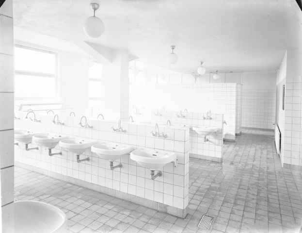 Schwarz-weiß-Foto einer öffentlichen Toilette mit Waschbecken, Hähnen, Wänden, Lampen und Fenstern.