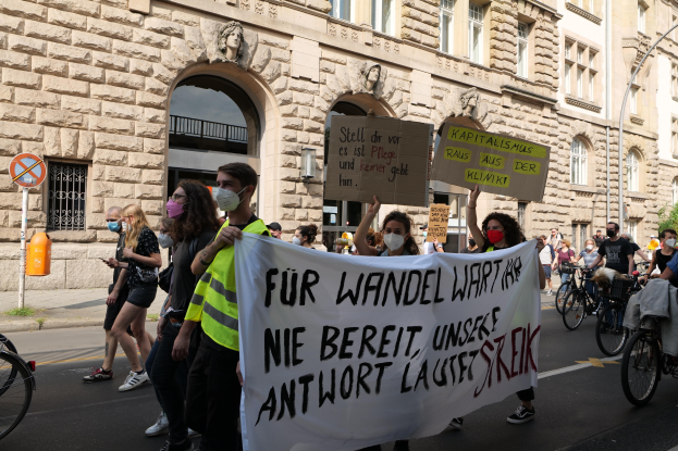 Eine Gruppe von Menschen marschiert auf einer Straße bei einer Demonstration, einige halten Schilder und andere fahren Fahrräder, mit einem Gebäude im Hintergrund, das Fenster, Bogengänge, Pfeiler und Skulpturen aufweist.