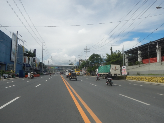 Eine belebte Stadtstraße mit zahlreichen Fahrzeugen, Gebäuden, Bäumen, einer Brücke und einem bewölkten Himmel im Hintergrund.