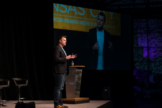 Eine Person in schwarzem Hemd spricht auf einem Seminar, neben einem Tisch mit einer Flasche, einem Stuhl dahinter, einem Projektor, der links von ihnen Inhalte anzeigt, und einem schwarzen Vorhang im Hintergrund.