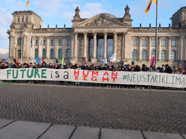 Eine Gruppe von Menschen h"alt eine Schilder mit der Aufschrift "Zukunft ist ein Mensch" vor dem Reichstaggeb"ude in Berlin, Deutschland, das mit Fenstern, S"ulen, Bogeng"angen und Statuen verziert ist und von Fahnen mit St"angen umgeben ist, unter einem bew"olkten Himmel.