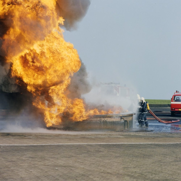 Ein Feuerwehrauto steht in Flammen am Straßenrand, zwei Personen mit Helmen halten Schläuche in der Nähe und ein Fahrzeug sowie der Himmel sind im Hintergrund zu sehen.