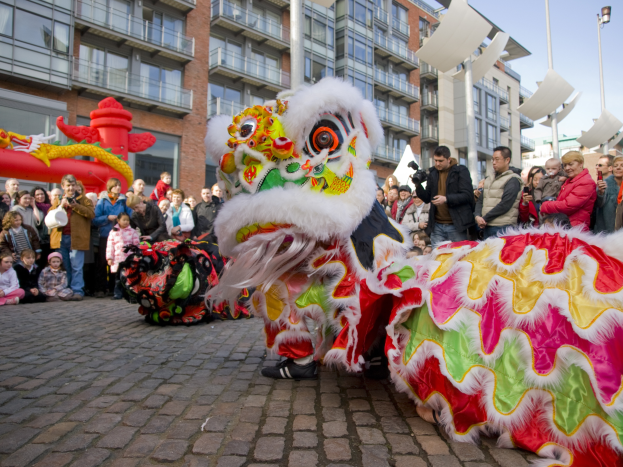 Eine Löwen-Tanzvorstellung während eines chinesischen Neujahrsfestes in Amsterdam, mit einer Zuschauermenge und Menschen, die den Moment mit Kameras festhalten, vor einem Hintergrund aus Gebäuden und einem klaren blauen Himmel.
