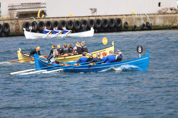 Eine Gruppe von Menschen in einem blauen und gelben Boot auf dem Wasser, die Paddel halten, mit einer Wand mit Reifen und einem Gebäude im Hintergrund, was ein Regatta vermuten lässt.