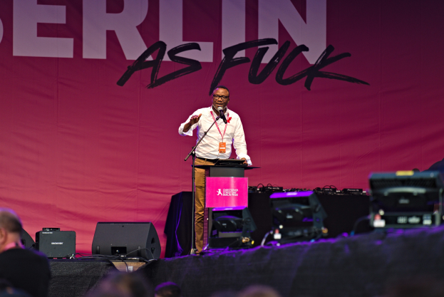 Mann in weißem Hemd und braunen Hosen, der in ein Mikrofon spricht, vor einem pinken "Berlin as Fuck"-Plakat auf der Bühne, vor einem Publikum.