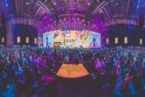 Eine große Menschenmenge sitzt auf Stühlen vor einer hell erleuchteten Bühne mit mehreren stehenden Personen, wahrscheinlich auf einem Online-Marketing-Rockstars-Event.