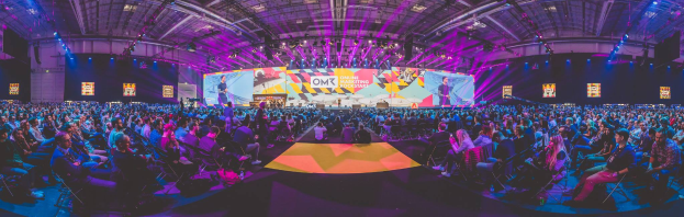 Eine große Menschenmenge sitzt auf Stühlen vor einer hell erleuchteten Bühne mit mehreren stehenden Personen, wahrscheinlich auf einem Online-Marketing-Rockstars-Event.