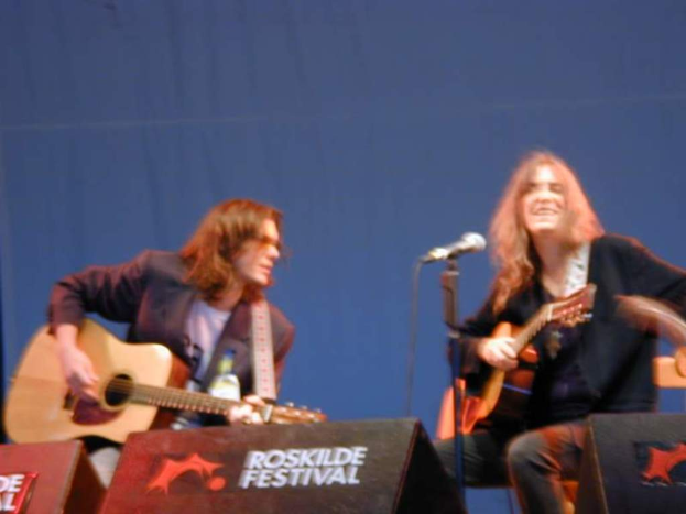 Zwei Männer auf der Bühne mit Musikinstrumenten, ein Mikrofon davor und zwei Kisten mit "ROCK SLIDE FESTIVAL" darauf.