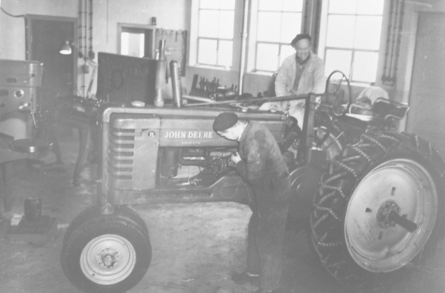 Zwei Männer arbeiten an einem John Deere-Traktor in einer Fabrik, einer sitzt auf dem Traktor und der andere steht daneben, mit Fenstern im Hintergrund, dargestellt in Schwarz-Weiß.