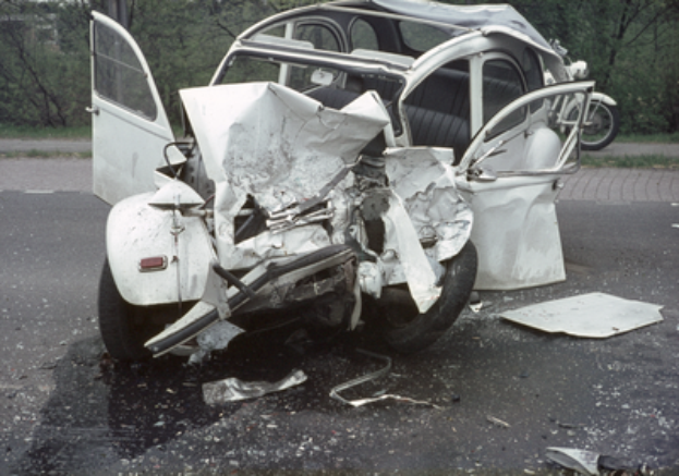 Ein Auto mit schwerer Frontalbeschädigung steht am Straßenrand, mit Bäumen, einem Gebäude und einem anderen Fahrzeug im Hintergrund.