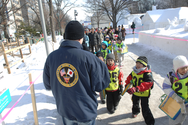 Eine Gruppe von Kindern geht eine schneebedeckte Straße entlang, mit einem Mann in einer blauen Jacke und Mütze im Vordergrund, gesäumt von Bäumen, Pfählen und Gebäuden, unter einem sichtbaren Himmel.