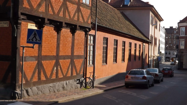 Fahrzeuge auf der Straße mit Gebäuden, Laternen, Verkehrszeichenmasten, Fenstern und Dächern im Hintergrund und einem Fahrrad auf dem Gehweg neben einem Mast.