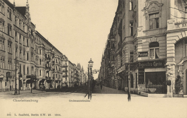 Altes Schwarz-Weiß-Foto einer Stadtstraße in Charlottenburg, Deutschland, mit Gebäuden, Straßenlaternen, Schildern, Motorfahrzeugen, Fußgängern, Gittern und einem klaren Himmel.