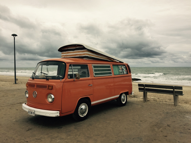 Oranger VW-Bus auf einem Strand in Strandn├Ąhe geparkt, mit einer Bank und einem Laternenmast in der N├Ąhe, unter einem bew├Âlktem Himmel mit dem Wasser im Hintergrund.