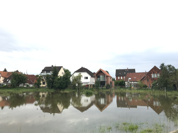 Eine überflutete Straße mit Häusern im Hintergrund, umgeben von Wasser, Gras, Pflanzen, Bäumen und Pfählen, unter einem sichtbaren Himmel, der die verheerenden Auswirkungen von Überschwemmungen in Deutschland verdeutlicht.