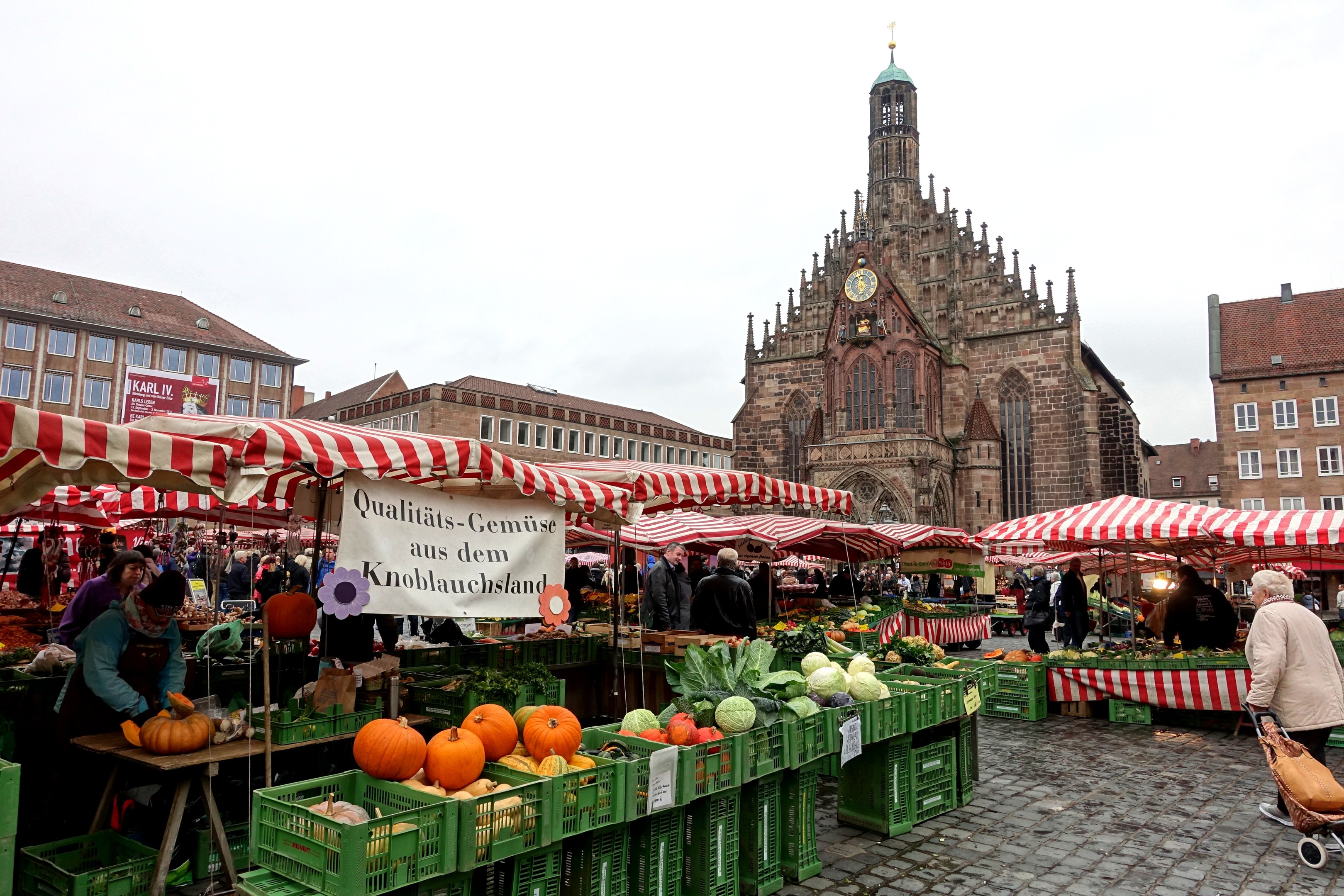 Ein belebter Markt in Nürnberg, Deutschland, mit Ständen, auf denen verschiedene Obst- und Gemüsesorten angeboten werden, Menschen, die einkaufen, und Zelte im Hintergrund, mit Gebäuden und einem Uhrenturm im Hintergrund, unter einem klaren Himmel.