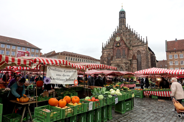 Ein belebter Markt in Nürnberg, Deutschland, mit Ständen, auf denen verschiedene Obst- und Gemüsesorten angeboten werden, Menschen, die einkaufen, und Zelte im Hintergrund, mit Gebäuden und einem Uhrenturm im Hintergrund, unter einem klaren Himmel.
