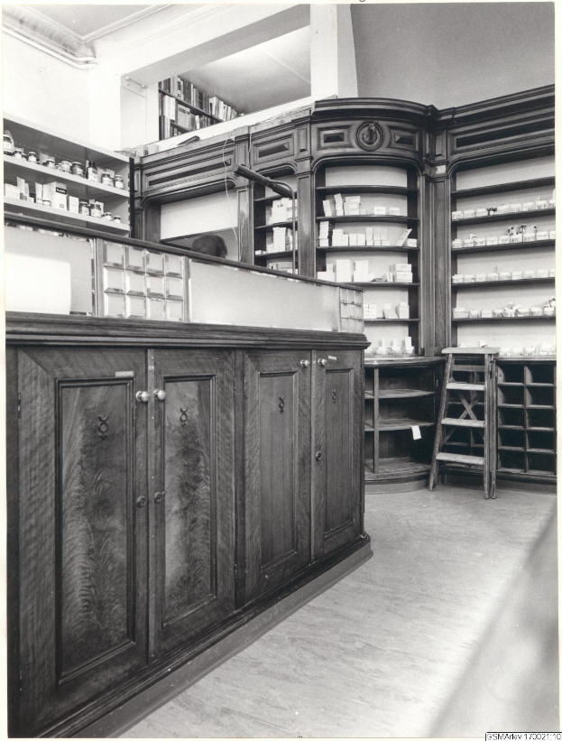 Ein Schwarz-Weiß-Foto eines Apothekeninneren mit einem Tresen, Schränken, einer Leiter und Regalen, die mit verschiedenen Gegenständen gefüllt sind, mit Text oben.