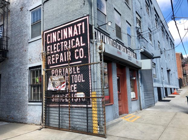 Das Geb├Ąude der Cincinnati Electrical Repair Co. mit Fenstern, T├╝ren, einer Schautafel, einem Metallzaun, einem M├╝llcontainer, Masten, Kabeln, B├Ąumen und einem bew├Âlktem Himmel.
