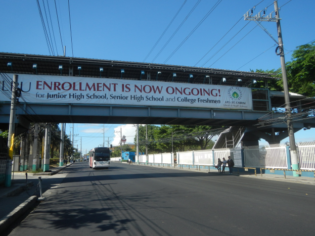Ein Bus fährt unter einer Brücke mit einem "Jetzt Anmelden"-Schild, Menschen gehen auf dem Gehweg darunter, Strommasten mit Drähten, Bäume und ein klarer blauer Himmel im Hintergrund.
