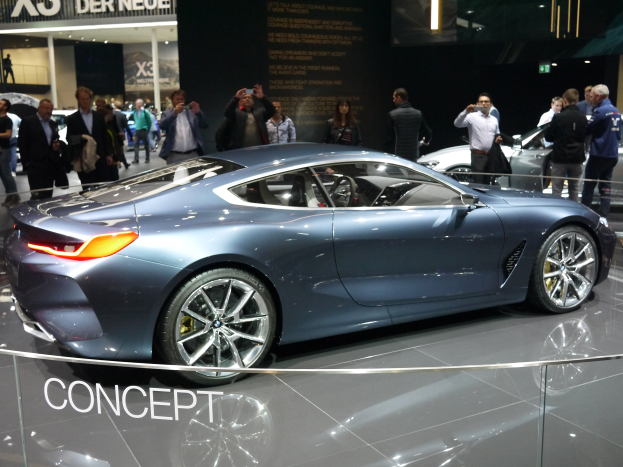 Ein BMW 8er-Konzeptfahrzeug mit glänzendem Schwarz und Chromakzenten wird auf der IAA in Frankfurt präsentiert, umgeben von Menschen, die Fotos machen, mit einem hell beleuchteten Hintergrund und einem Textschild im Bild.
