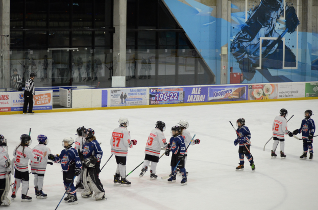 Junge Menschen in Helmen und Eishockeyausrüstung spielen Eis-hockey auf einer Indoor-Eisfläche mit Bannern, Säulen und Glaswänden im Hintergrund.