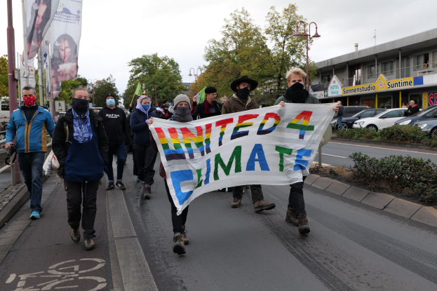Eine Gruppe von Menschen mit Masken geht eine Straße entlang und hält ein "United 4 Climate"-Schild hoch, mit einem Gehweg, Geländer, Schildern, Bäumen, Laternen, Fahrzeugen, Gebäuden und einem klaren blauen Himmel im Hintergrund.