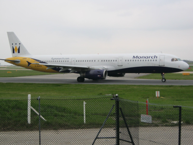Ein Monarch Airlines Airbus A320-200 auf der Rollbahn, umgeben von Gras und einem Zaun mit Pfählen und Brettern, mit Bäumen und einem klaren blauen Himmel im Hintergrund.