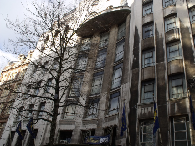 Gebäude mit Fenstern, einer Fahne und Flaggen an Masten unten sowie einem Baum mit Zweigen.