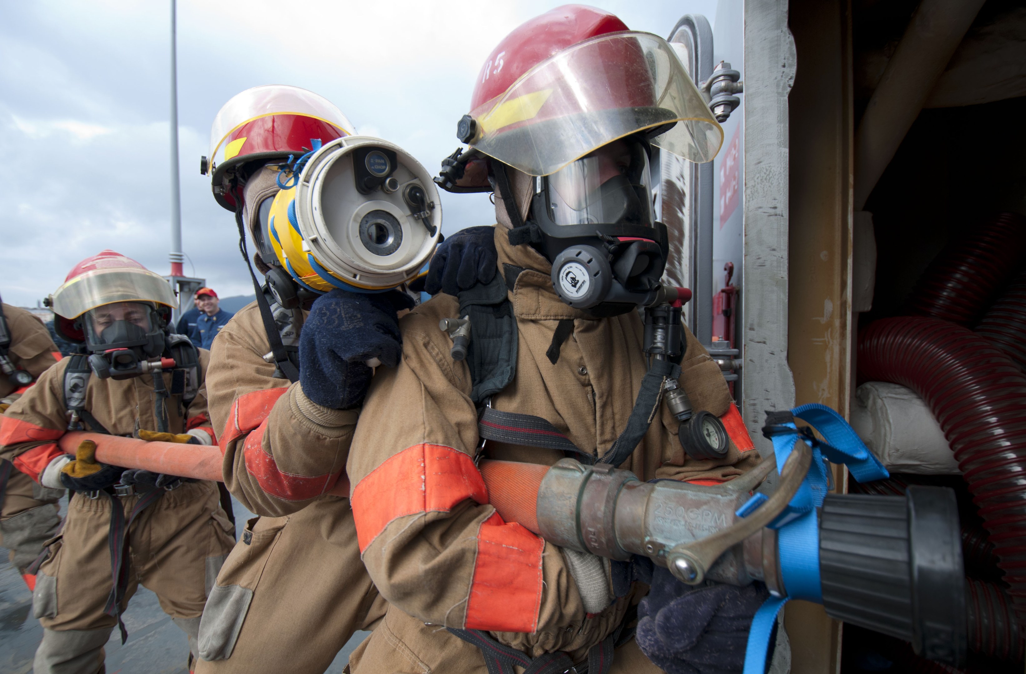 Feuerwehrleute in Schutzausrüstung mit einem Schlauch haltenden, einem Stab, bewölktem Himmel und Rohren auf der rechten Seite.