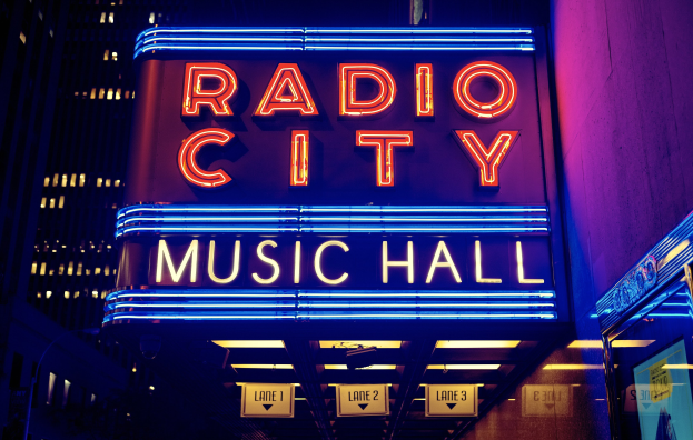 Hell erleuchtetes Radio City Music Hall in New York City mit umliegenden Hochhäusern und einem "Radio City"-Schild sowie mehreren kleineren Schildern an der Basis.