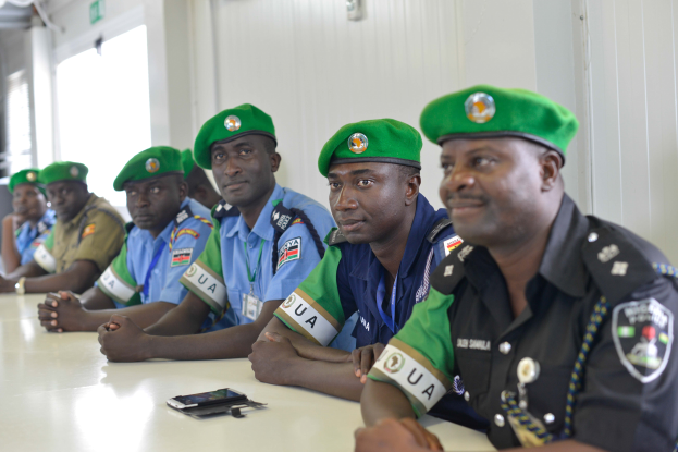 Eine Gruppe von Polizeibeamten sitzt an einem Tisch mit Mobiltelefonen und anderen Gegenständen in einem Raum mit einer Wand, Fenstern links und einer beleuchteten Decke.