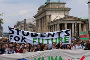 Eine Gruppe von Schülern marschiert in Berlin, hält eine bunt bemalte Fahne mit der Aufschrift "Schüler für die Zukunft", mit Gebäuden, Bäumen und Himmel im Hintergrund.