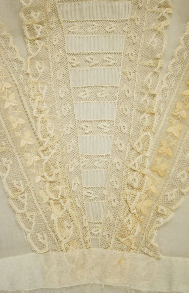 Nahaufnahme eines weißen Kleides mit zarten, detailreichen Spitzenverzierungen in verschiedenen Formen und Mustern auf einem leichten, luftigen Stoff.