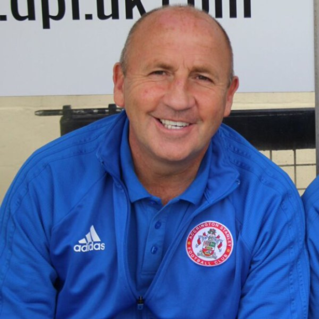 Zwei lachende Männer in einem Raum, einer in einer blauen Jacke, wahrscheinlich der Trainer des Accrington Stanley Football Club, mit einer Tafel an der Wand dahinter.